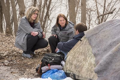 Two women talk with homeless man seating on the ground in front of a tent in the woods.